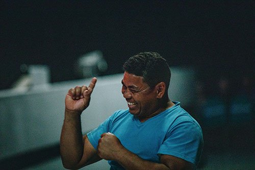 A man in a blue shirt is smiling broadly, raising his right hand with a playful gesture. The dimly lit and slightly blurred background highlights his joyful expression, reminiscent of the warmth and inclusivity web accessibility aims to bring to every user. - Clio Websites | Top Canadian Web Design Company