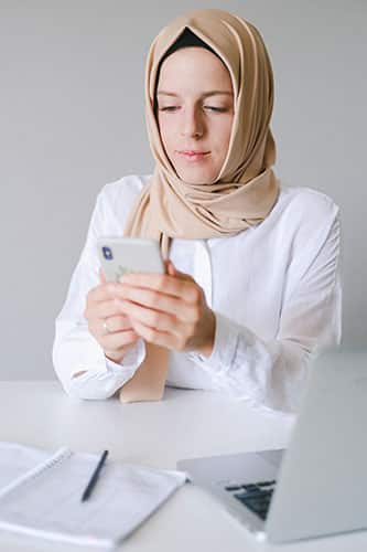 A woman wearing a beige hijab and white shirt sits at a desk, looking at her smartphone. A notebook, pen, and laptop—open to web design for medical offices—are on the white desk in front of her. - Clio Websites | Top Canadian Web Design Company
