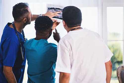 Three healthcare professionals in scrubs closely examine and discuss an X-ray image together in a bright room with large windows, highlighting the importance of teamwork and precision, much like effective web design for medical offices. - Clio Websites | Top Canadian Web Design Company
