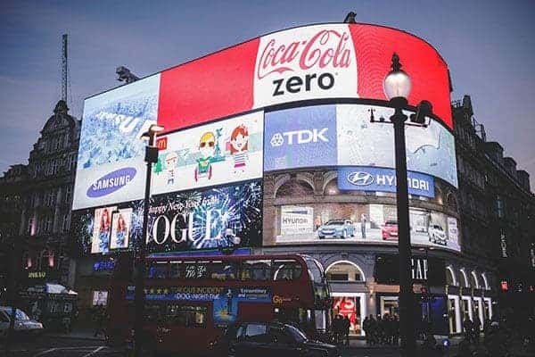 A busy city scene at dusk shows large electronic billboards promoting a website alongside ads for brands like Coca-Cola, Samsung, and Hyundai above a red double-decker bus and streetlights. - Clio Websites | Top Canadian Web Design Company