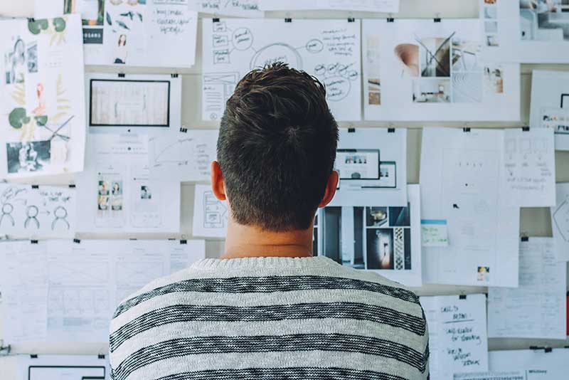 A person with short brown hair wearing a striped sweater stands before a wall covered in various papers, diagrams, and photos, reminiscent of a small business landing page. The scene suggests brainstorming or planning, with charts and notes meticulously attached to the wall. - Clio Websites | Top Canadian Web Design Company