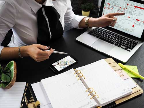 A person in business attire sits at a desk with an open planner, a laptop displaying a real estate web design floor plan, a smartphone, documents, a pen, and glasses, appearing to work and point at the screen. - Clio Websites | Top Canadian Web Design Company