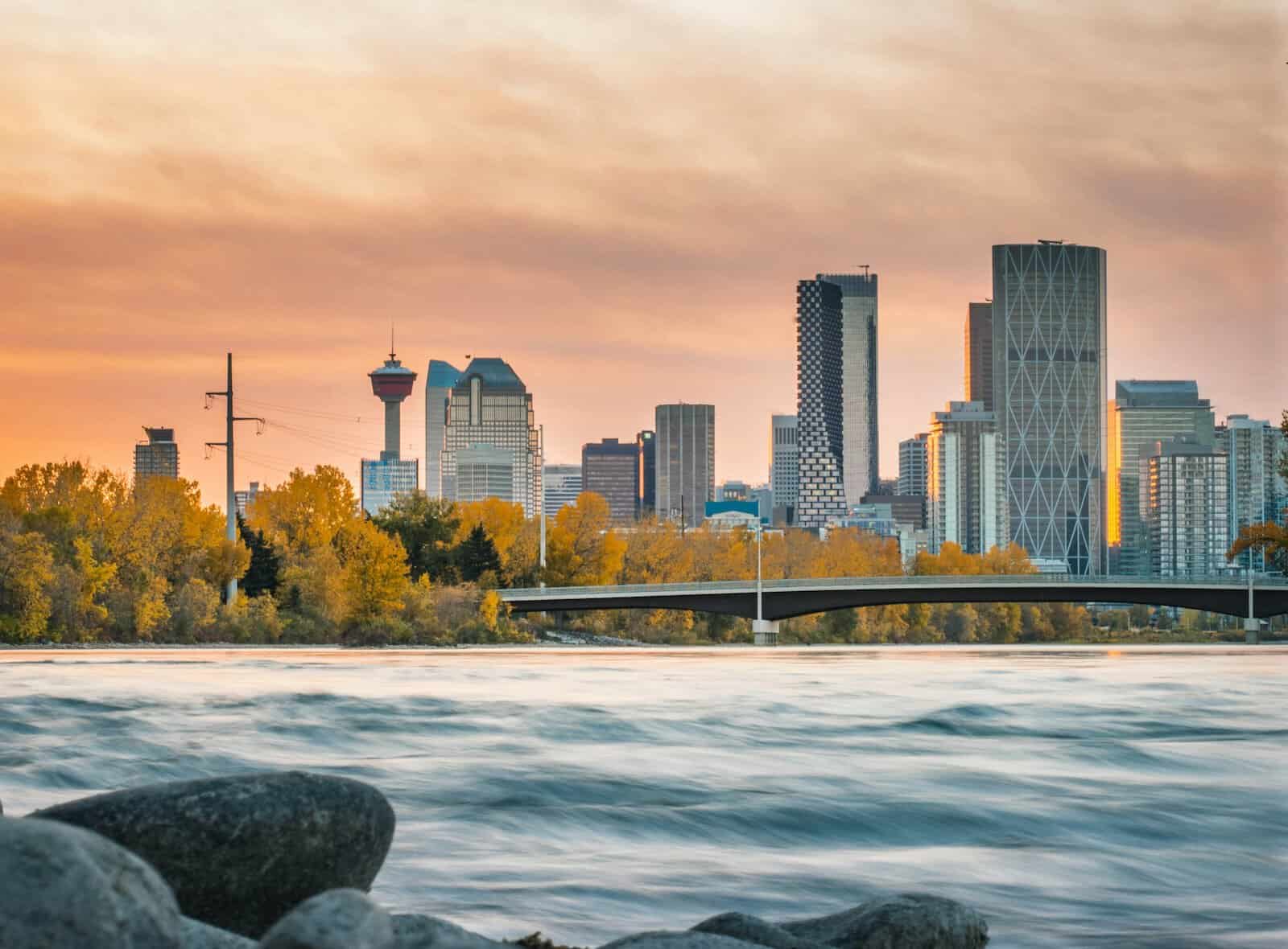 A city skyline during sunset with a river in the foreground. Skyscrapers and tall buildings, like those in Calgary, line the horizon, while golden autumn trees border the water's edge. A bridge spans the river, under a sky painted with warm hues, much like a Calgary web design masterpiece. - Clio Websites | calgary-web-design-calgary-skyline