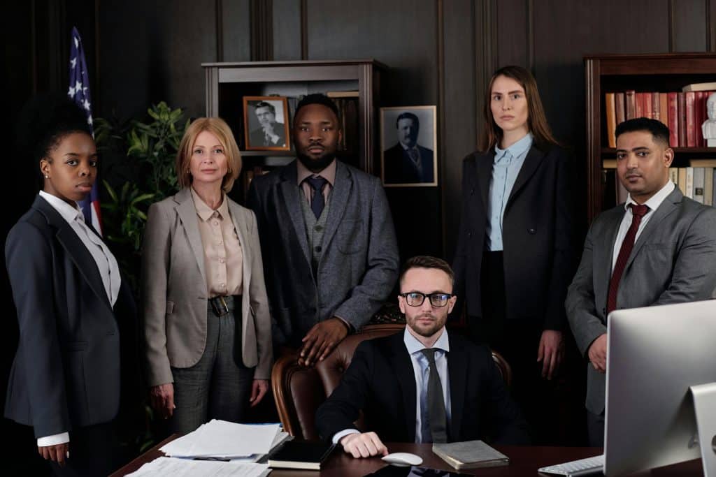 A group of five professionals in business attire gather in a formal office with bookshelves, documents, and a computer, conveying a serious work meeting—perfect for illustrating web design for family lawyer services. - Clio Websites | Top Canadian Web Design Company