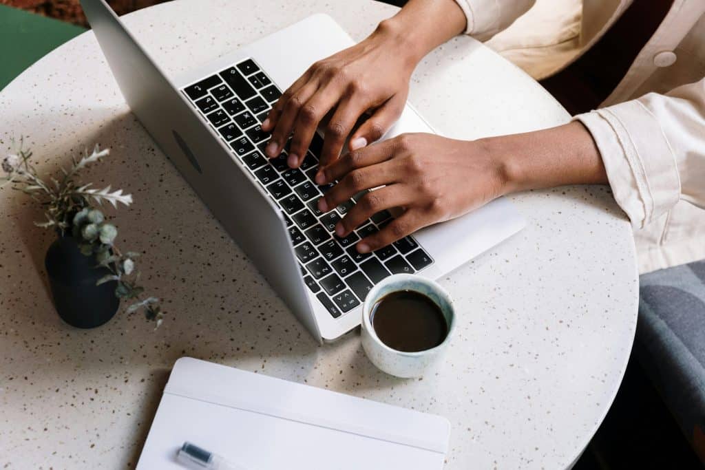 A person types on a laptop at a round, speckled table, meticulously building content. Beside them are a small plant, a cup of coffee, and a closed notebook with a pen. The light-colored shirt they wear complements the serene setting perfectly. - Clio Websites | Top Canadian Web Design Company