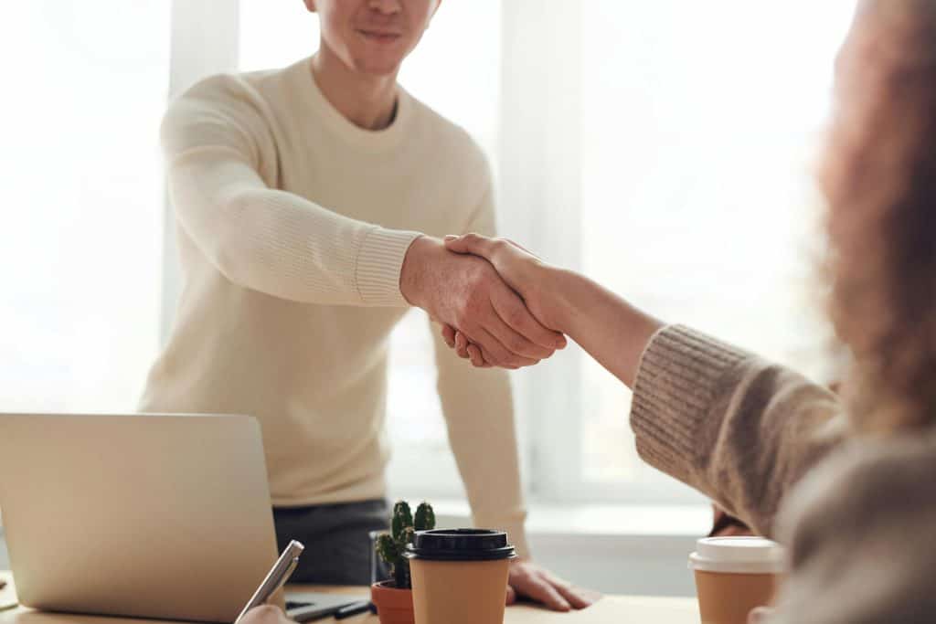 Two people shake hands over a desk with a laptop computer on it, symbolizing building trust with high-quality photos.