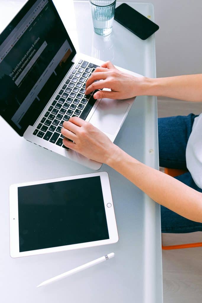 A person types on a laptop at a white desk with a tablet, stylus, smartphone, and glass of water nearby, suggesting a modern workspace ideal for web design for photographers. - Clio Websites | Top Canadian Web Design Company