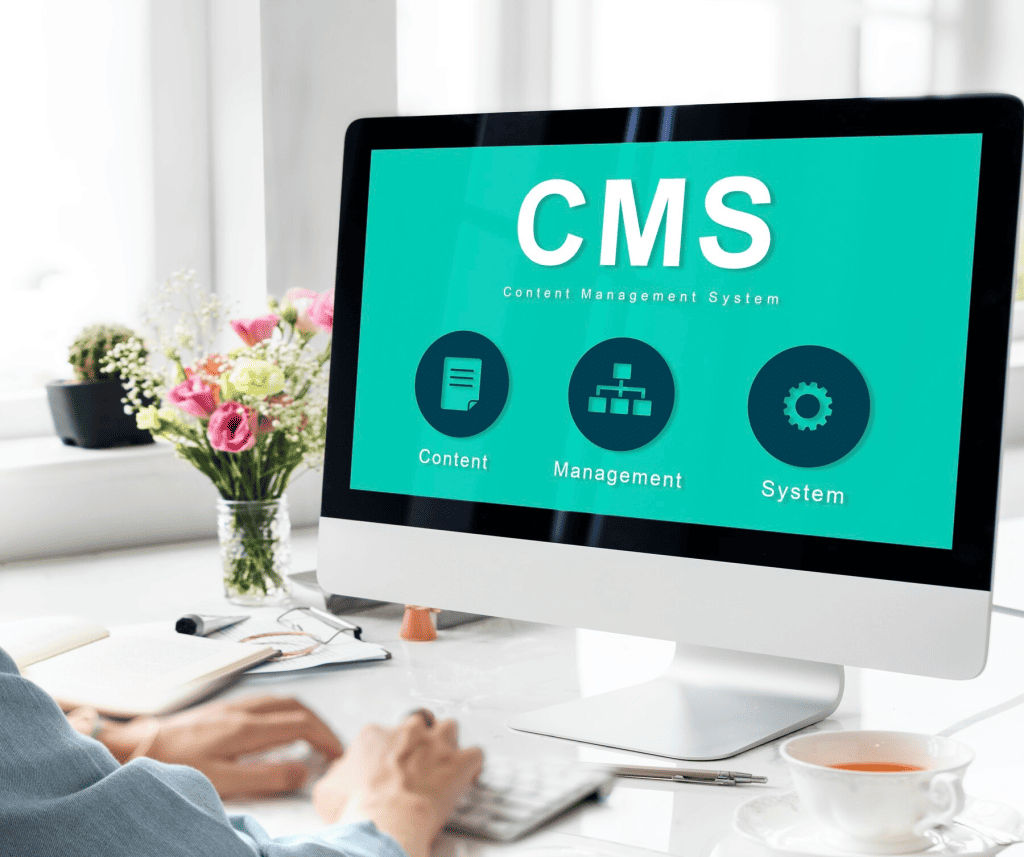 A business owner is working on a desktop computer displaying a CMS screen, essential for managing website costs efficiently. The monitor shows icons for content and management. The workspace includes a plant, flowers, and stationery on a pristine white desk. - Clio Websites | Top Canadian Web Design Company