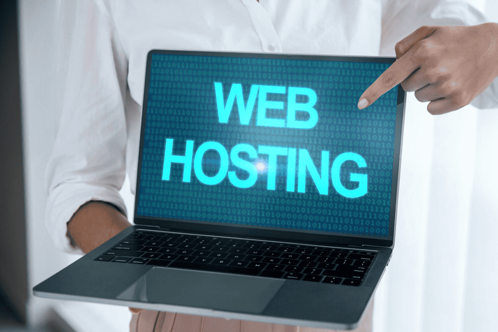 A person is holding a laptop displaying the words "WEB HOSTING" in large letters across the screen. A business owner is pointing at the display, emphasizing the text. The background is blurred, focusing all attention on how essential hosting is for eCommerce success. - Clio Websites | Top Canadian Web Design Company