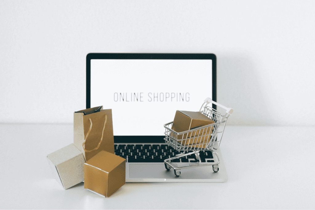 A laptop displaying the words "Online Shopping" on its screen sits behind a small shopping cart filled with boxes and two paper shopping bags, perfectly capturing the essence of eCommerce. It's an ideal setup for any business owner looking to explore the digital marketplace. - Clio Websites | Top Canadian Web Design Company