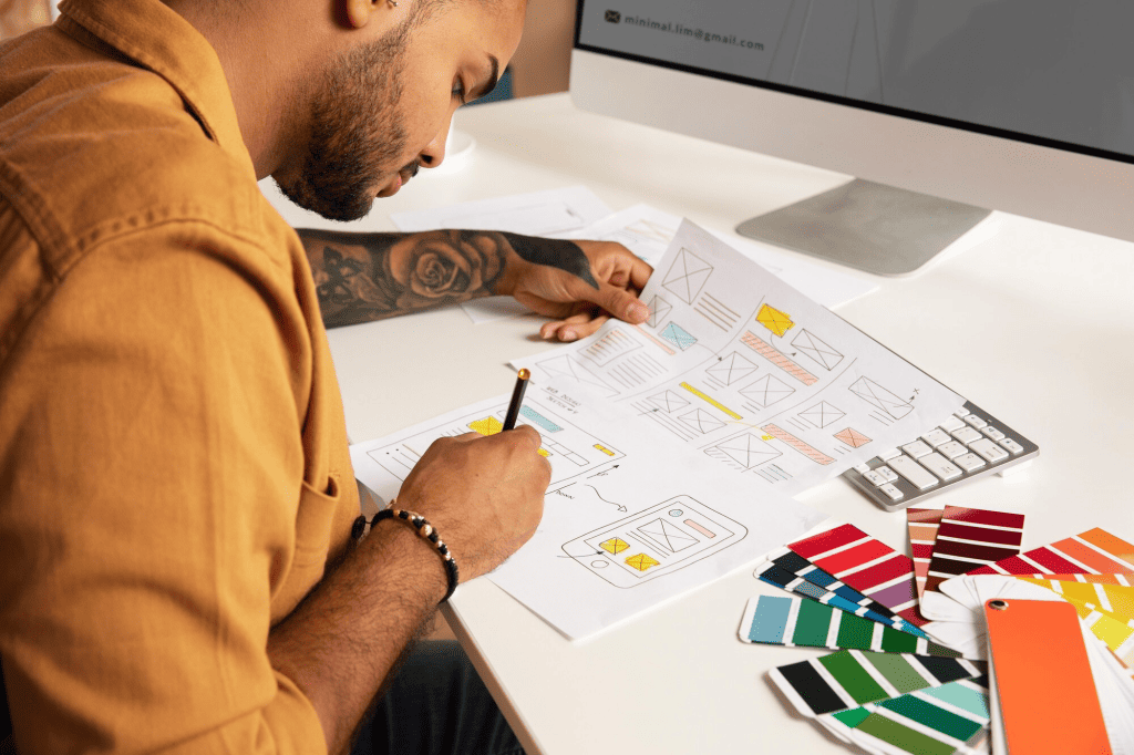 A person in a brown shirt is drawing website designs on paper at a desk, with a color palette and computer in the background. Various design sketches and color swatches are scattered on the table. - Clio Websites | Top Canadian Web Design Company
