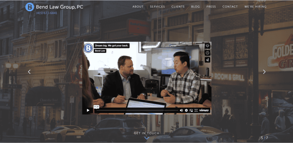 A video from Bend Law Group, PC, showcasing two men in discussion within an office setting. The background prominently features images of city streets and a menu typical of law firm websites, with options like About, Services, Clients, and more. - Clio Websites | Top Canadian Web Design Company