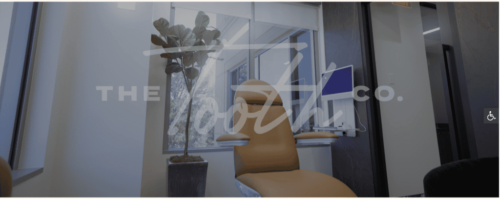 A modern dentist office with a tan dental chair facing a large window, exemplifying exquisite dental design. A potted plant rests beside the chair, and a monitor is mounted on the wall. The words "The Tooth Co." are subtly displayed on the glass, reflecting top-tier aesthetic considerations. - Clio Websites | Top Canadian Web Design Company