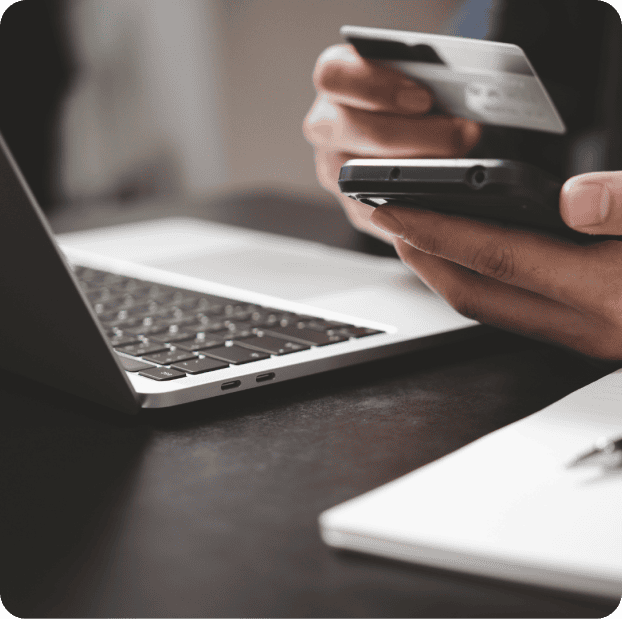 Close-up of a person holding a credit card and using a smartphone, with a laptop and notebook on a dark table. The scene illustrates modern ecommerce development, suggesting online shopping or mobile banking in today's digital workspace. - Clio Websites | Top Canadian Web Design Company