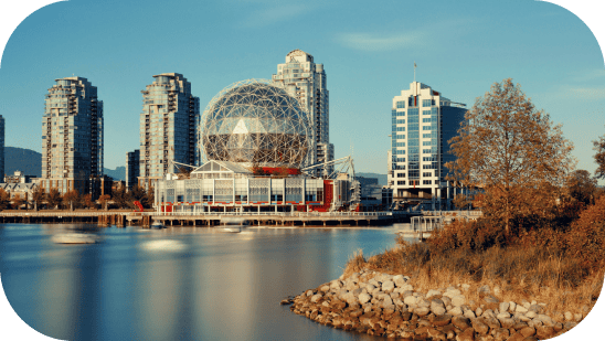 A scenic view of Montreal's modern city waterfront showcases a large geodesic dome surrounded by high-rise buildings. The calm water reflects the clear blue sky, with autumn foliage and a rocky shoreline in the foreground—a perfect backdrop for inspiring web design concepts. - Clio Websites | Top Canadian Web Design Company