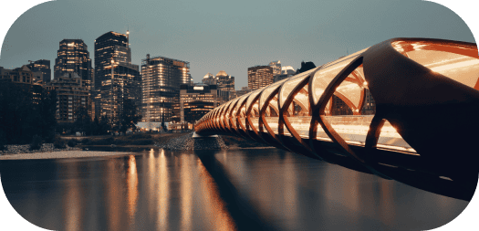 A cityscape at dusk showcases a uniquely designed illuminated bridge spanning over water, embodying the artistry akin to Calgary web design. The tube-like structure with bright lights contrasts against a backdrop of modern buildings with their lights glowing in the evening sky. - Clio Websites | Top Canadian Web Design Company