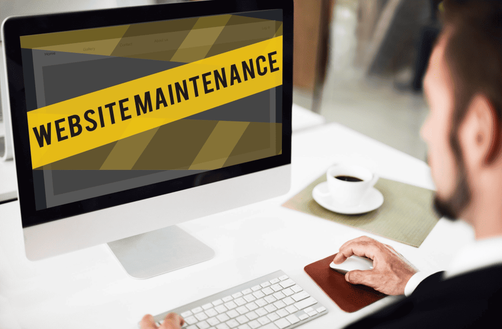 A business owner in a suit sits at a desk working on a computer. The monitor displays a diagonal yellow and black striped banner with the text "Website Maintenance." A cup of coffee is on the desk beside him as he considers eCommerce strategies and website costs. - Clio Websites | Top Canadian Web Design Company