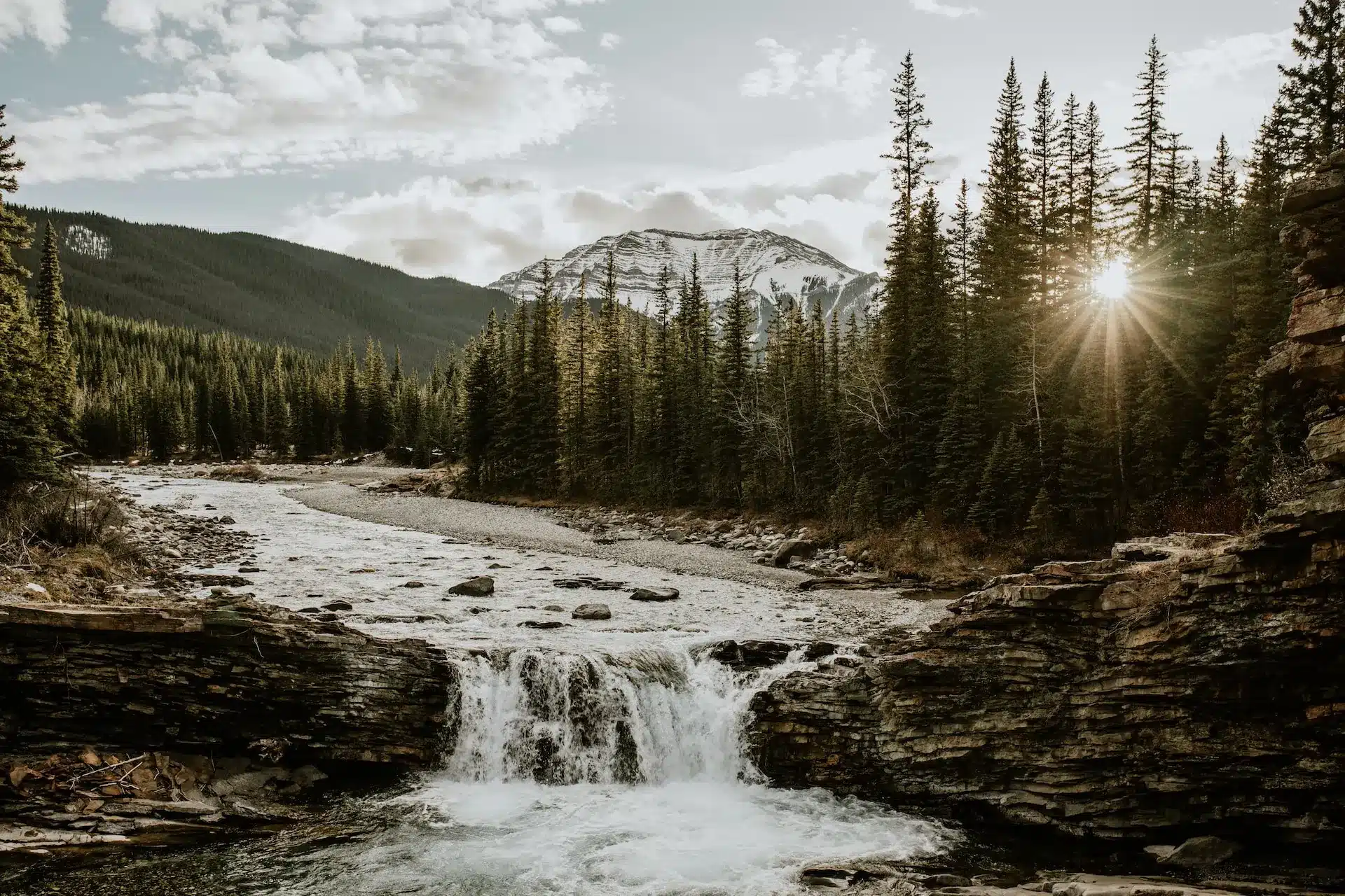 A serene Alberta landscape features a river cascading over rocks in the foreground. Evergreen trees line the river, with a majestic snow-capped mountain in the background. The sun peeks through the trees, casting a warm glow reminiscent of elegant web design artistry. - Clio Websites | Alberta Web Design Company