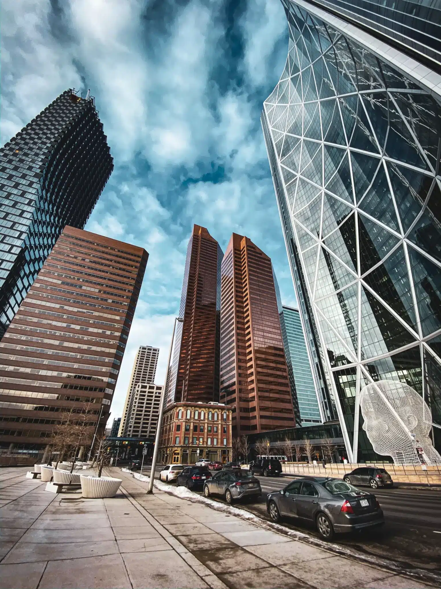 A cityscape featuring modern skyscrapers under a cloudy sky. Prominent buildings include a sleek glass structure reminiscent of Calgary web design with geometric patterns and brick high-rises. Cars are parked along the street lined with planters and a small historic building. - Clio Websites | Top Canadian Web Design Company