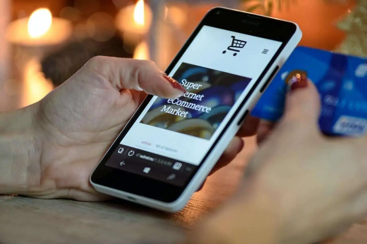 A person is holding a smartphone displaying an e-commerce website, showcasing the results of savvy ecommerce development. In the other hand is a blue credit card, suggesting online shopping or payment activities. The background is softly blurred with warm lighting, adding to the scene's inviting allure. - Clio Websites | Top Canadian Web Design Company