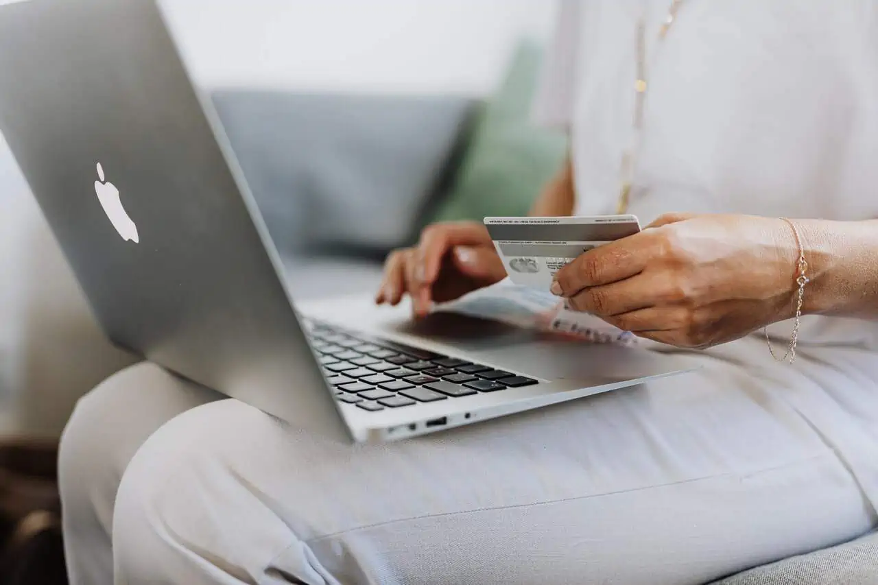 A person sits on a couch holding a credit card while using a silver laptop, showcasing the seamless ease of modern ecommerce development. Dressed in white clothing, they immerse themselves in online shopping or banking activities. - Clio Websites | Top Canadian Web Design Company