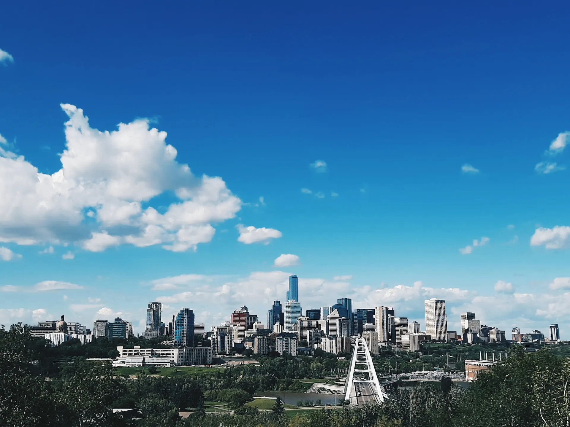 A vibrant city skyline under a bright blue sky with scattered clouds showcases tall skyscrapers surrounded by lush greenery. A distinctive bridge structure completes the scene, reminiscent of Edmonton's innovative web design blending nature and architecture seamlessly. - Clio Websites | Top Canadian Web Design Company