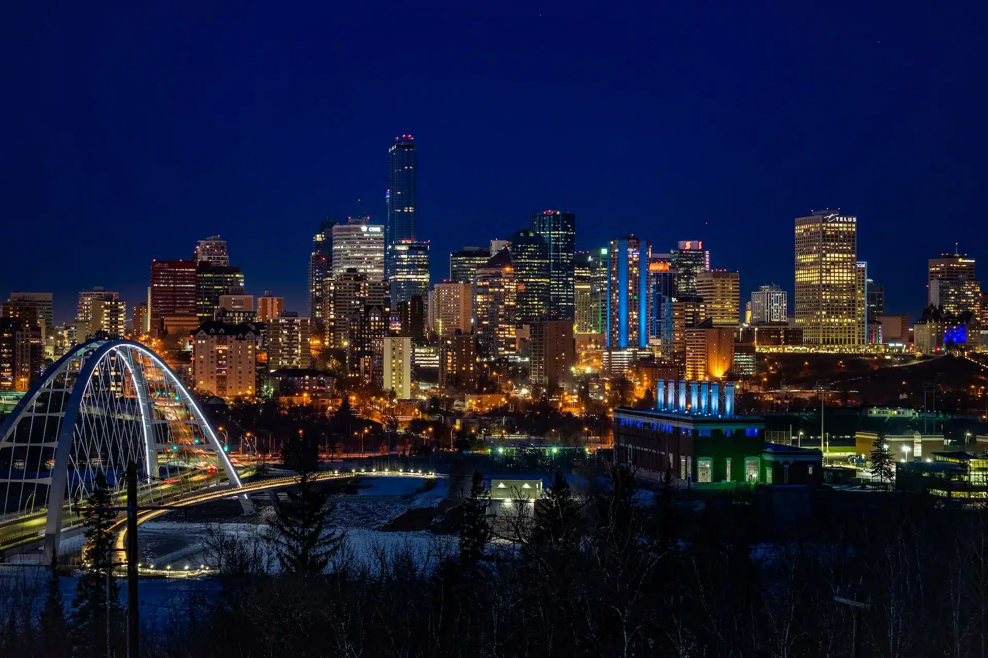 A vibrant nighttime cityscape featuring Edmonton's skyline of illuminated skyscrapers against a deep blue sky. In the foreground, a bridge is lit up, with a river and trees visible below—mirroring the seamless flow of creative design intertwined in Edmonton's web design landscape. - Clio Websites | Top Canadian Web Design Company