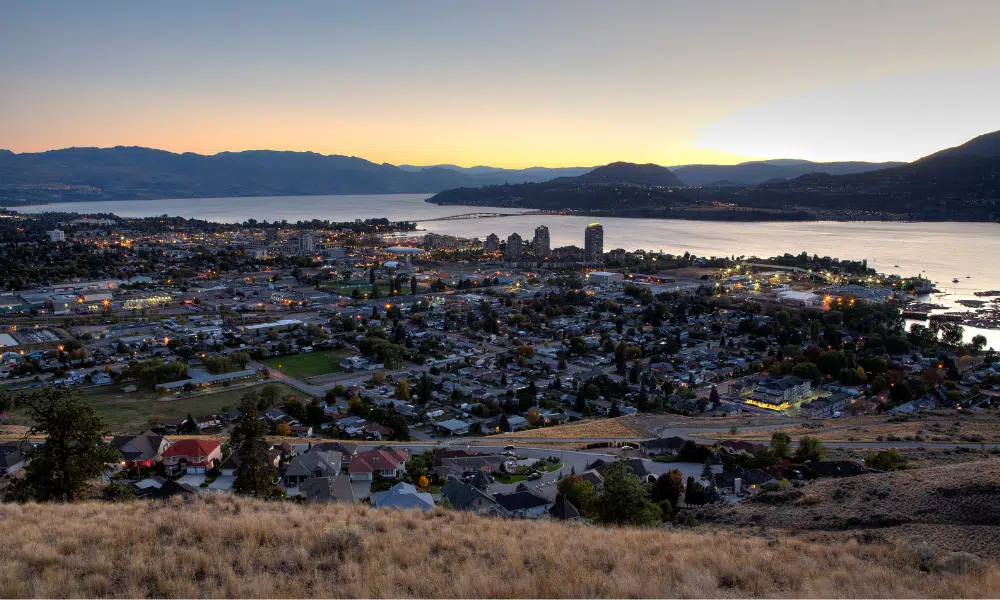 Panoramic view of Kelowna at dusk with hills in the foreground and a large lake. The city, known for its dynamic web design scene, is lit by streetlights and surrounded by mountains under a clear sky, with the horizon glowing from the setting sun. - Clio Websites | Top Canadian Web Design Company