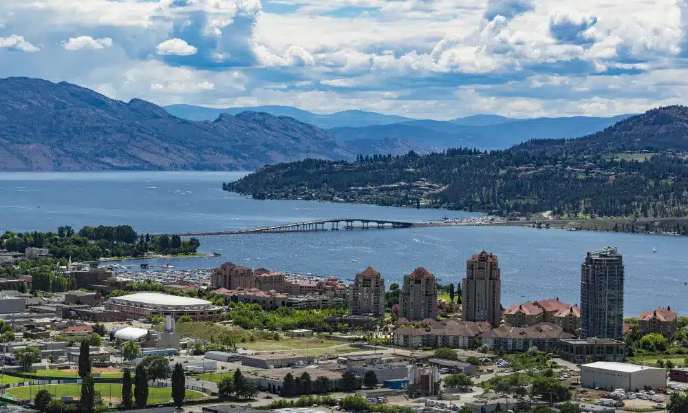 A picturesque view of a lakeside city, reminiscent of Kelowna, with a bridge gracefully extending over the water, surrounded by lush green hills and distant mountains under a partly cloudy sky. Buildings and greenery blend seamlessly in the foreground, much like an expertly crafted web design. - Clio Websites | Top Canadian Web Design Company