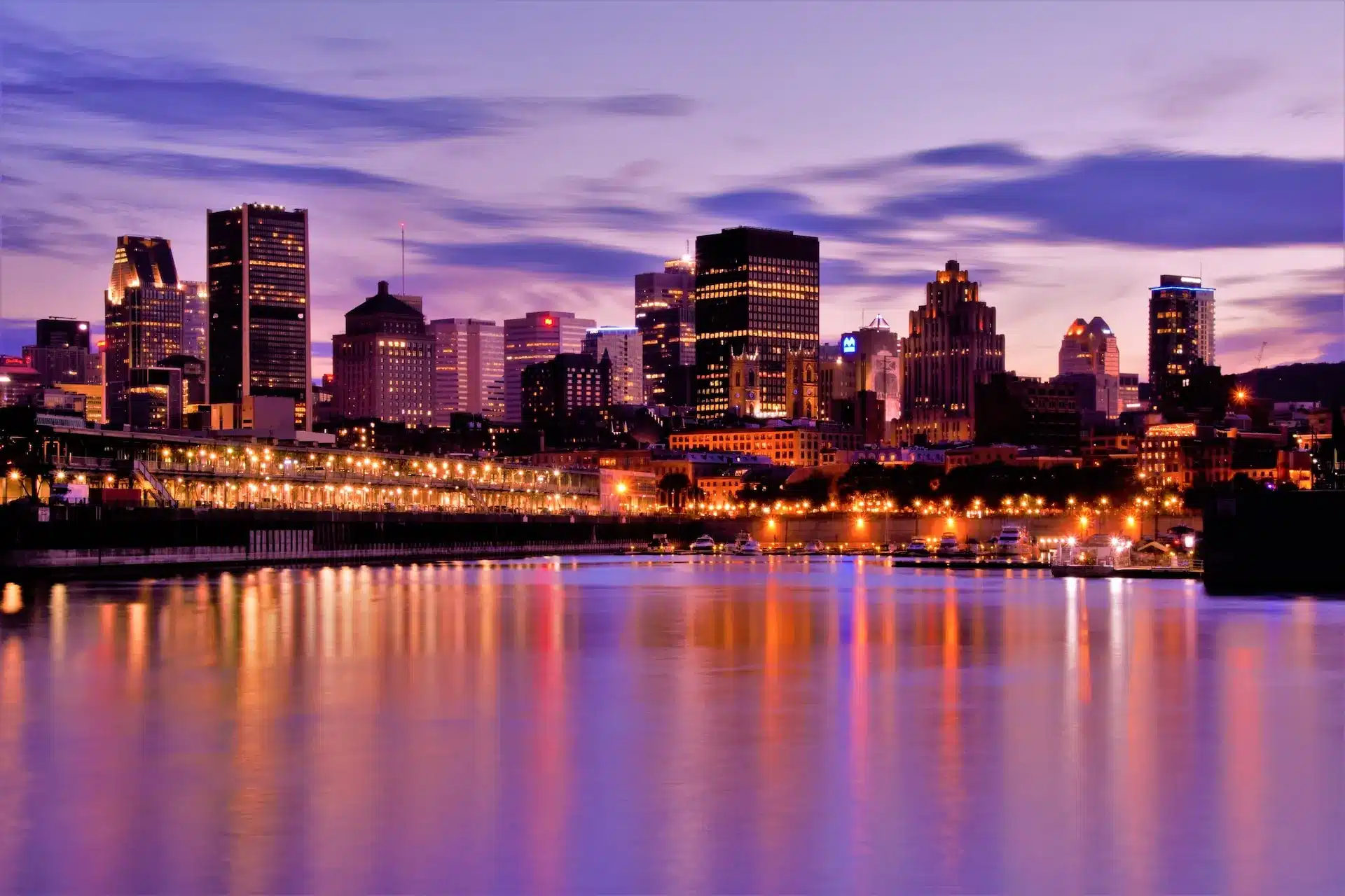 A city skyline at dusk with tall buildings illuminated against a purple and blue sky, reminiscent of Montreal's unique charm. The lights from the city reflect on the calm water in the foreground, adding a vibrant glow to the scene, much like an artist crafting web design brilliance. - Clio Websites | Top Canadian Web Design Company