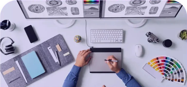 A person uses a graphics tablet on a tidy desk with dual monitors displaying design sketches, a keyboard, a sketchpad, a camera, headphones, and a color wheel. The workspace is well-organized and creative. - Clio Websites | Top Canadian Web Design Company