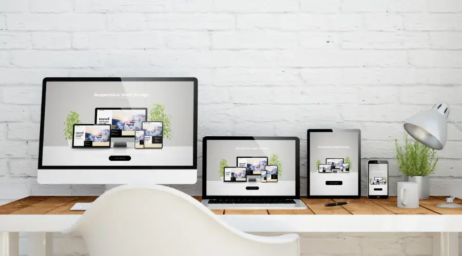 A desk with a computer, laptop, tablet, and smartphone, all displaying a website with the text "Responsive Web Design." A white chair is in front of the desk, with a gray lamp and a potted plant beside the devices against a white brick wall. - Clio Websites | Top Canadian Web Design Company