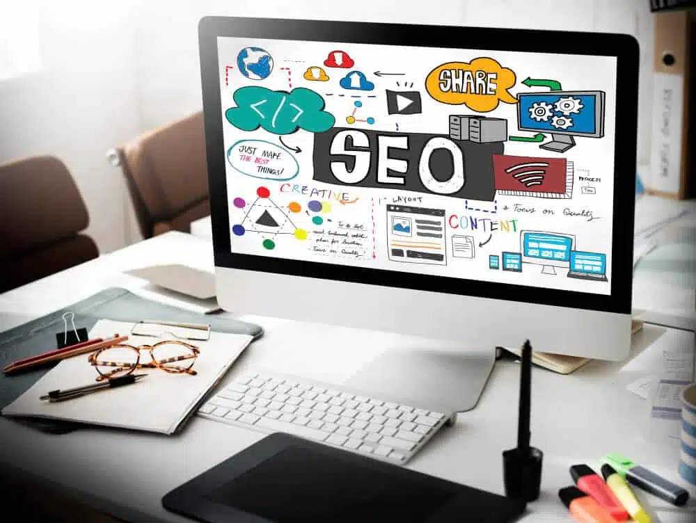 A computer screen displaying a colorful digital collage related to SEO, showcasing our services with icons, graphs, and text. The desk in front has open notebooks, a smartphone, a graphics tablet, and a pen. Bright office setting with sunlight streaming in. - Clio Websites | Top Canadian Web Design Company