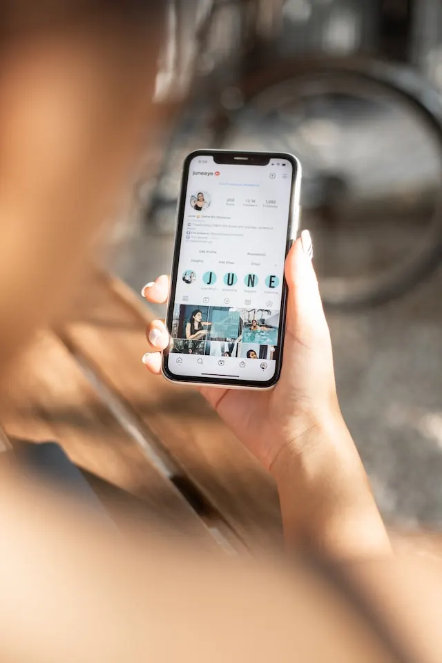 A person is holding a smartphone that displays an Instagram profile with a clean pfp. The screen shows a grid of photos beneath the profile header. The scene is set outdoors, with blurred elements softly blending into the background. - Clio Websites | Top Canadian Web Design Company