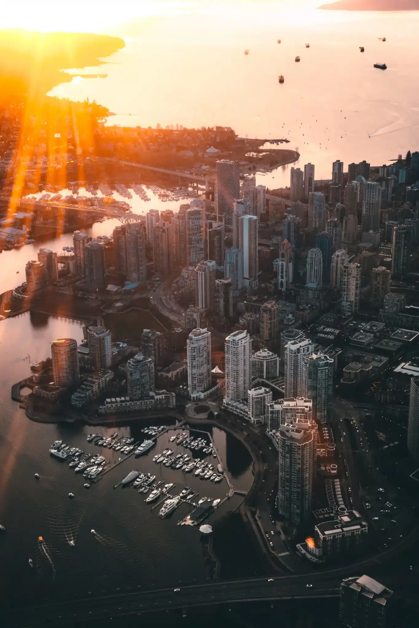 Aerial view of Vancouver's cityscape at sunset, showcasing tall buildings and a marina with boats surrounded by tranquil water. The sun casts a warm, golden light, creating dramatic shadows akin to masterful web design highlights across the vibrant scene. - Clio Websites | Top Canadian Web Design Company