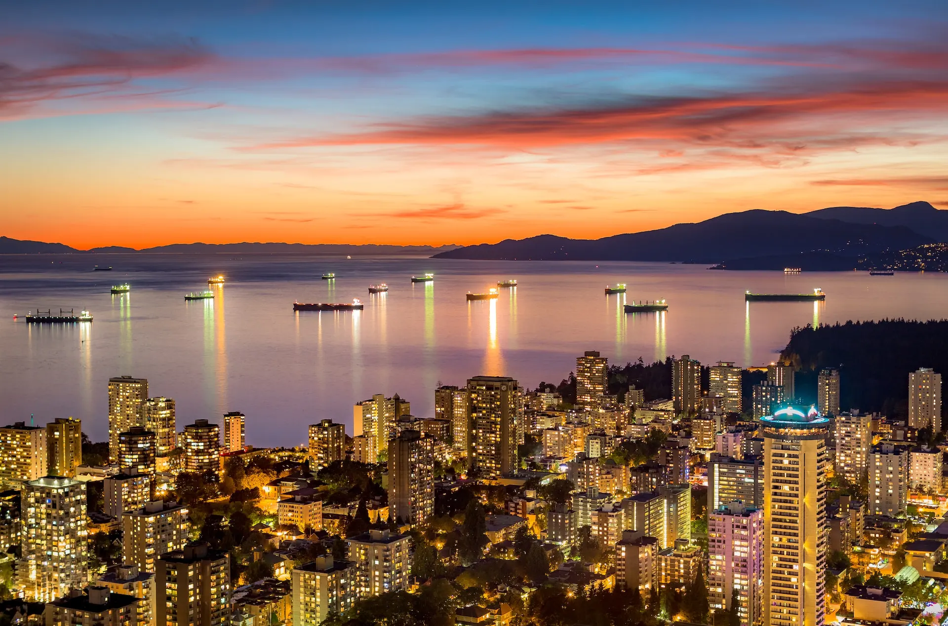 A stunning view of Vancouver's cityscape at sunset, with illuminated buildings that echo the creativity of web design in the foreground. The vibrant orange and pink hues reflect on the calm water, where several ships are anchored, while distant mountains silhouette the background. - Clio Websites | Top Canadian Web Design Company
