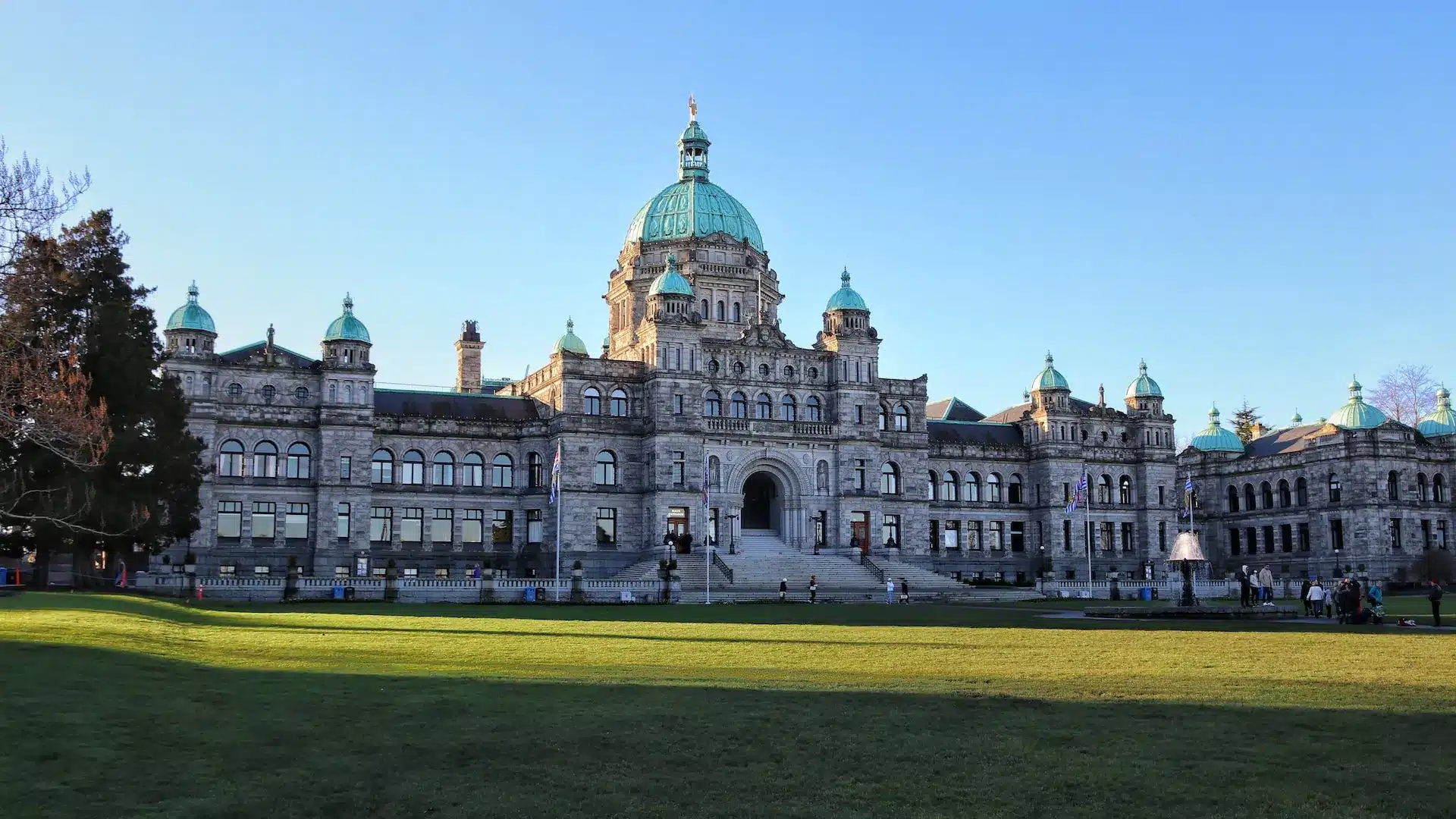 The image captures a grand, historic government building with a central dome and smaller green-capped towers. Set against a clear blue sky, the scene includes a well-kept lawn and people strolling by. Much like Victoria's approach to web design, it blends elegance with functionality seamlessly. - Clio Websites | Top Canadian Web Design Company