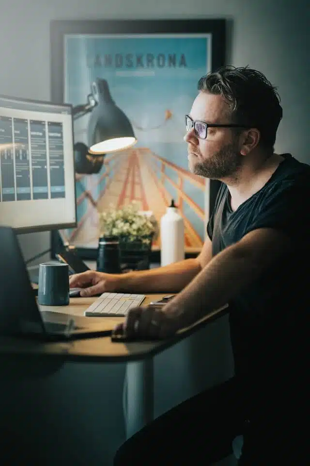 A person with glasses sits at a desk, intently focusing on Calgary webflow design projects displayed across multiple computer windows. The room is dimly lit, with a desk lamp illuminating a framed picture. Nearby, there’s a mug and a water bottle, essentials for the creative process. - Clio Websites | Top Canadian Web Design Company