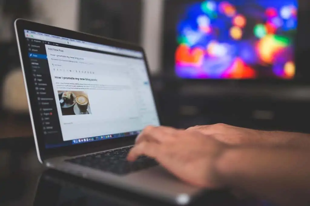 A person types on a laptop, juggling website maintenance as a blogging platform lies open. The screen displays a draft post with text and an image of a coffee cup, while the background is blurred, revealing the glow of a colorful television screen. - Clio Websites | Top Canadian Web Design Company