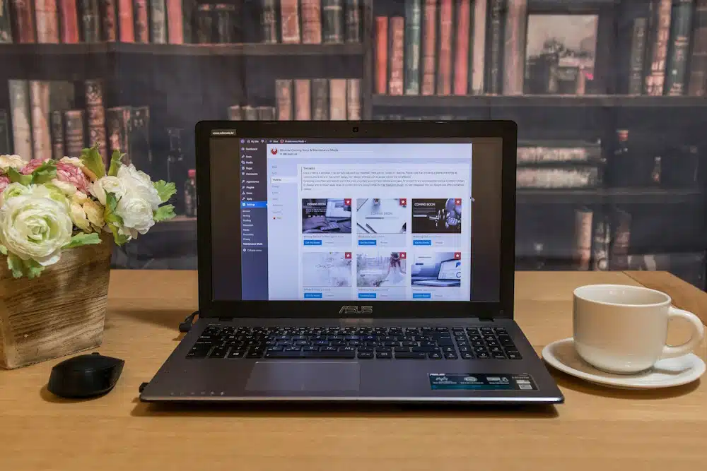 A laptop displaying a WordPress dashboard sits on a wooden table in Calgary. Beside it, a potted plant with white and pink flowers, a black computer mouse, and a white cup and saucer add charm. In the background, a developer’s bookshelf filled with old books completes the scene. - Clio Websites | Top Canadian Web Design Company