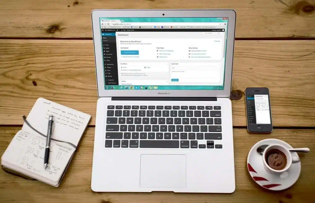 A laptop showing the WordPress dashboard rests on a wooden table, illustrating the ease of accessing our services. Nearby lie an open notebook with a pen, a smartphone, and a steaming cup of coffee. - Clio Websites | Top Canadian Web Design Company
